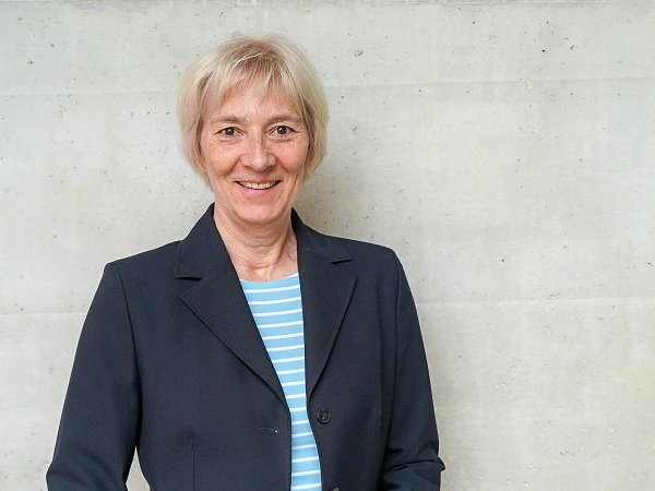 Die Frau mit blonden, kurzen Haaren trägt einen schwarzen Blazer über einem gestreiften Shirt und lächelt vor einer neutralen, grauen Wand.
