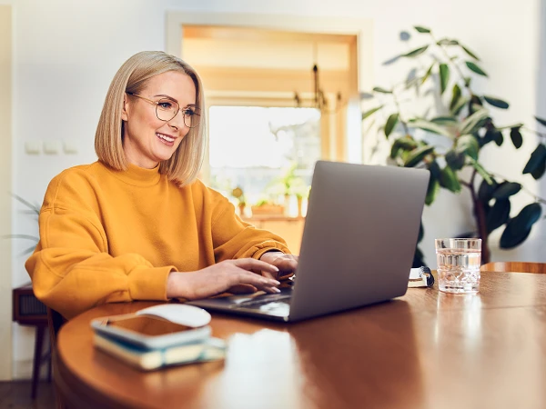 Eine lächelnde Frau mit blonden, schulterlangen Haaren sitzt an einem Holztisch und arbeitet am Laptop. Sie trägt einen orangefarbenen Pullover und eine Brille. Im Hintergrund sind Pflanzen und ein helles Raumambiente zu sehen. Ein Wasserglas und ein Notizbuch liegen auf dem Tisch.