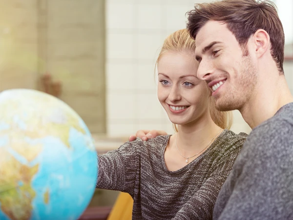 Ein lächelndes Paar sitzt eng beieinander und schaut auf einen Globus. Die Frau berührt mit einer Hand eine Stelle auf dem Globus, während der Mann sie aufmerksam ansieht.
