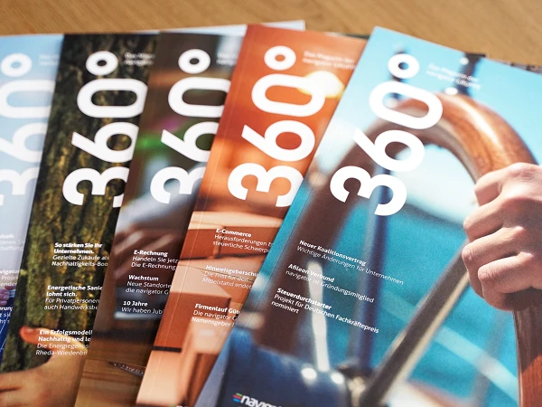 Eine Reihe von achtseitigen Magazinen mit dem Titel "360°", die auf einem Tisch angeordnet sind. Die Cover zeigen verschiedene Farben und eine größere Schriftart. Im Hintergrund sind unscharfe Details einer Hand zu sehen, die ein Steuerrad hält.