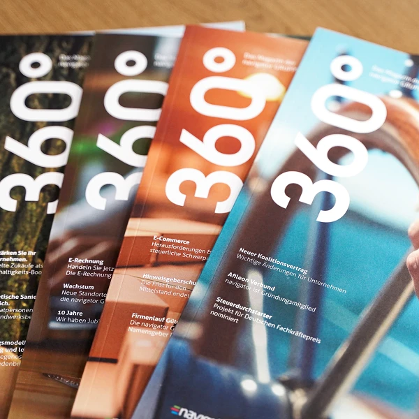 Eine Reihe von achtseitigen Magazinen mit dem Titel "360°", die auf einem Tisch angeordnet sind. Die Cover zeigen verschiedene Farben und eine größere Schriftart. Im Hintergrund sind unscharfe Details einer Hand zu sehen, die ein Steuerrad hält.