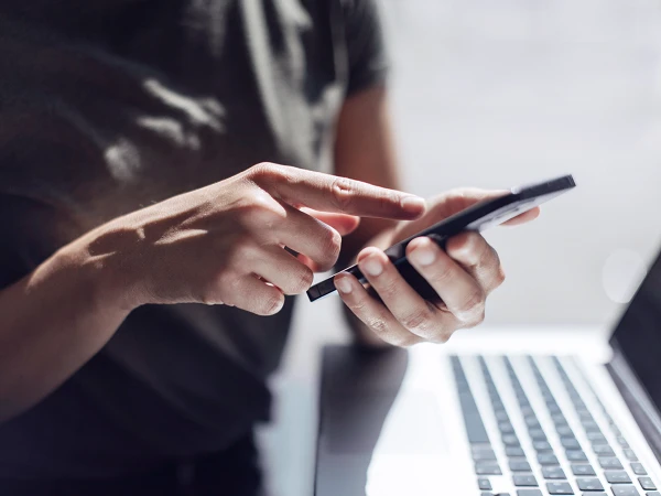 Eine Person in einem grauen T-Shirt nutzt ein Smartphone, während sie mit der anderen Hand auf den Bildschirm tippt. Im Hintergrund ist ein Laptop sichtbar.