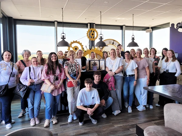 Eine Gruppe von etwa 25 Personen posiert in einem modernen Innenraum mit großen Fenstern und dekorativen Zahnrädern im Hintergrund. Einige halten kleine Gegenstände in der Hand, während sie lächeln und in die Kamera schauen.