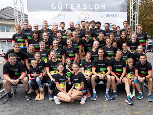 Eine große Gruppe von Menschen steht vor einem Banner mit der Aufschrift "Gütersloh", alle tragen einheitliche schwarze Shirts mit bunten Streifen. Die Gruppe besteht aus Männern, Frauen und Kindern, die fröhlich zusammen posieren, einige halten Medaillen oder zeigen ihre Startnummern.