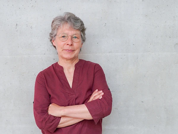 Eine Frau mit kurzen grauen Haaren und Brille steht mit verschränkten Armen vor einer grauen Betonwand, sie trägt ein rotes Oberteil und schaut direkt in die Kamera.