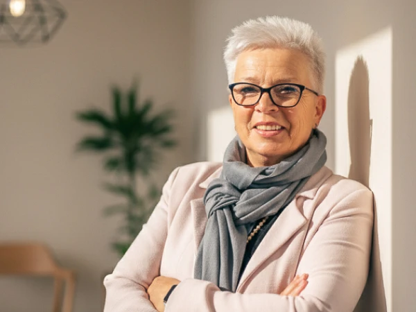 Eine lächelnde ältere Frau mit kurzen, grauen Haaren und einer Brille steht lässig mit verschränkten Armen da. Sie trägt einen hellen Blazer und einen grauen Schal, und im Hintergrund ist eine Pflanze sowie ein modernes Lichtobjekt sichtbar.