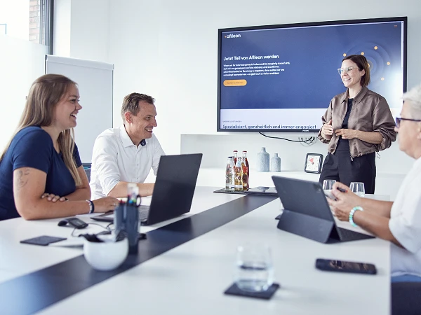 In einem modernen Besprechungsraum präsentieren sich vier Personen während eines Meetings, während eine Frau an der Wand ein digitales Präsentationsmaterial zeigt. Auf dem Tisch stehen Getränke und Laptops.