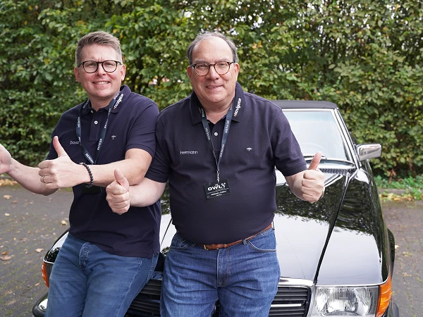 Zwei Männer in Polo-Shirts sitzen auf der Motorhaube eines schwarzen Autos und zeigen mit beiden Daumen nach oben. Im Hintergrund sind grüne Bäume zu sehen.