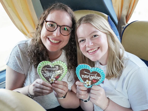 Zwei lächelnde Frauen halten Herzplätzchen in den Händen, die mit buntem Zuckerguss dekoriert sind, während sie in einem Bus sitzen.