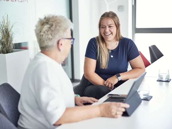 Zwei Personen sitzen an einem Tisch in einem hellen Büroraum und unterhalten sich. Eine Frau mit blonden, kurzen Haaren trägt eine weiße Jacke und schaut die andere Frau an, die lange, blonde Haare hat und in einem blauen Shirt gekleidet ist. Auf dem Tisch steht ein Tablet und ein Glas Wasser.