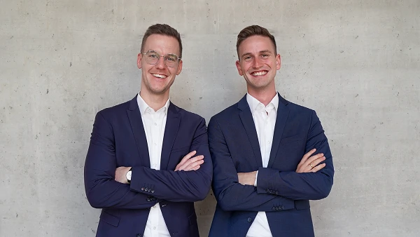 Zwei junge Männer stehen nebeneinander vor einer grauen Betonwand, beide tragen elegante Blazer und lächeln freundlich, während sie mit verschränkten Armen posieren.