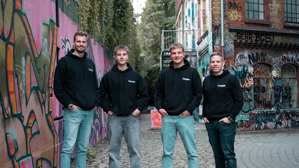 Vier Personen stehen in einer bunten Gasse, die mit Graffiti bedeckt ist. Sie tragen schwarze Sweatshirts und lässige Jeans, während sie locker nebeneinander stehen und in die Kamera schauen. Im Hintergrund sind weitere graffitibedeckte Wände und Pflanzen zu sehen.