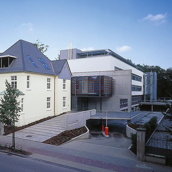 Moderne Architektur mit einem flachen Gebäude und einem historischen Haus in einer städtischen Umgebung, umgeben von Bäumen und Pflanzen.