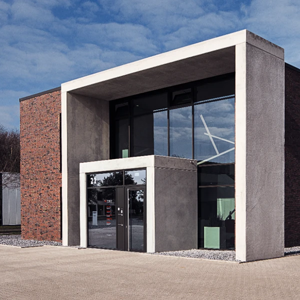Ein modernes Gebäude mit einer Kombination aus rotem Ziegel und Beton, featuring große Fenster und ein Vordach; der Platz vor dem Gebäude ist gepflastert.
