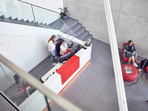 Eine moderne Empfangshalle mit einem Empfangstresen, an dem zwei Personen beschäftigt sind. Im Hintergrund sind graue Wände und eine Treppe zu sehen, während sich auf der linken Seite eine Person in einem roten Sessel mit einem Getränk entspannt.
