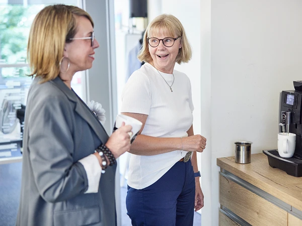 Zwei Frauen stehen in einem modernen Büro und unterhalten sich, während die eine Frau eine Tasse Kaffee hält und die andere an einer Kaffeemaschine steht und lächelt.