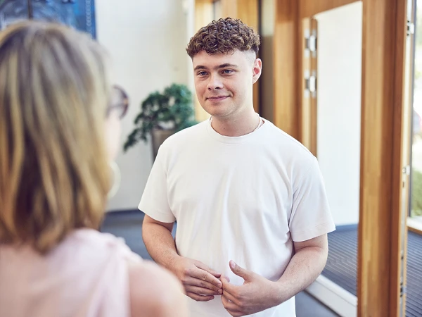 Ein junger Mann mit lockigem, blondem Haar trägt ein einfaches weißes T-Shirt und lächelt, während er mit einer Person spricht, die ihm gegenübersteht. Im Hintergrund sind Holz- und Glasdetails des Raumes zu sehen.