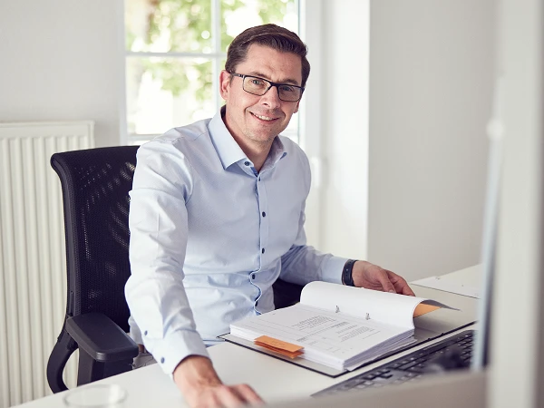 Ein Mann mit Brille sitzt an einem Schreibtisch und lächelt, während er auf ein geöffnetes Dokument blickt. Im Hintergrund sind ein Fenster und eine Heizung zu sehen.