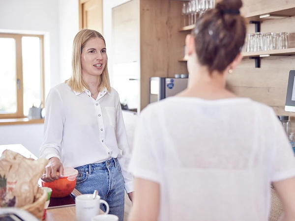 Zwei Frauen stehen in einer modernen Küche; eine Frau spricht mit einer Handbewegung, während die andere Frau ihr zuhört. Auf dem Tisch befinden sich verschiedene Utensilien und ein Glas.