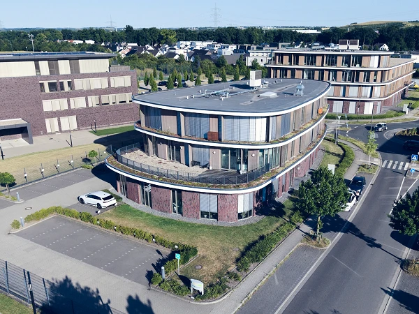 Eine moderne Gebäudestruktur mit geschwungener Fassade und großen Fenstern steht an einer Kreuzung, umgeben von gepflegten Grünflächen und Parkplätzen. Im Hintergrund sind weitere moderne Gebäude und ein Waldgebiet sichtbar.