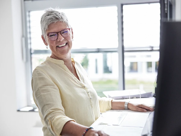 Eine lächelnde Frau mit kurzen grauen Haaren und Brille sitzt an einem Schreibtisch und schaut freundlich in die Kamera. Sie trägt ein hellbeiges Hemd und steht vor einem Computerbildschirm, während im Hintergrund Fenster mit Tageslicht zu sehen sind.