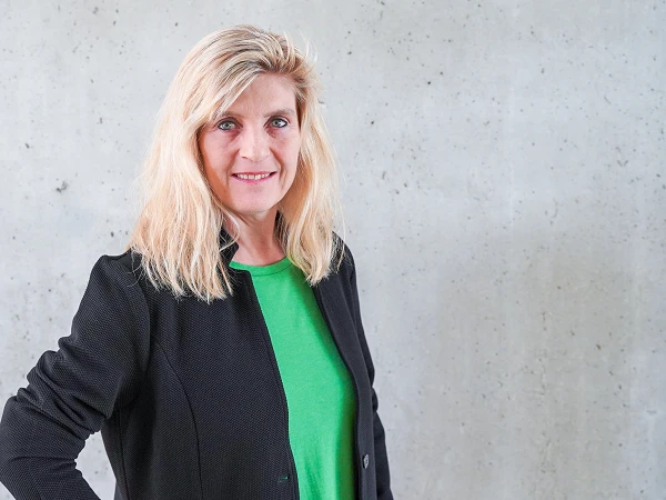 Eine Frau mit langen blonden Haaren trägt ein grünes Shirt und eine schwarze Jacke. Sie steht lächelnd vor einer grauen, strukturierten Wand.
