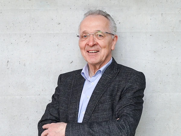 Ein älterer Mann mit grauen Haaren und einer Brille steht lächelnd vor einer grauen Betonwand und trägt einen dunklen Anzug.