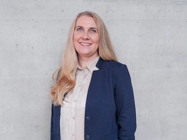 Eine blonde Frau mit langen, glatten Haaren lächelt vor einer ungefähren grauen Betonwand. Sie trägt ein helles Hemd und einen dunklen Blazer.
