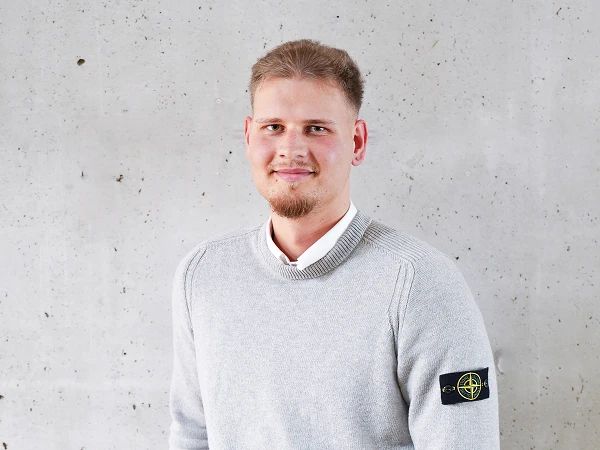 Ein junger Mann mit kurzen, blonden Haaren und einem leichten Bart trägt einen grau gestrickten Pullover mit einem Patch auf dem linken Ärmel. Er steht vor einer grauen Wand mit einer strukturierten Oberfläche und lächelt freundlich.