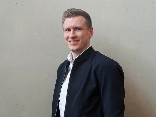 Eine Person mit kurzen Haaren und einem freundlichen Lächeln steht vor einer grauen Wand und trägt eine dunkle Jacke über einem weißen Hemd.