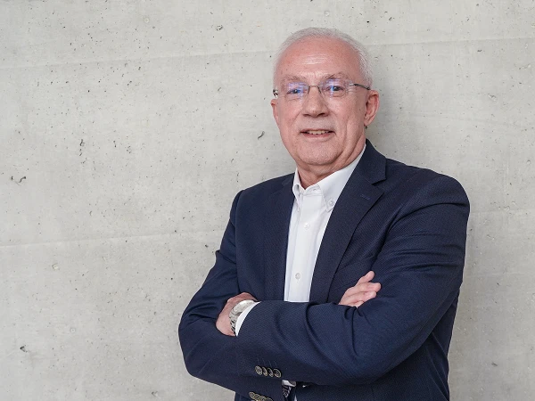 Ein älterer Mann mit grauen Haaren und Brille steht lächelnd mit verschränkten Armen vor einer grauen, strukturierten Wand. Er trägt ein dunkelblaues Sakko und ein weißes Hemd.