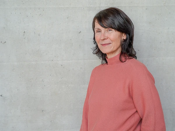 Eine lächelnde Frau mit langen, dunklen Haaren steht seitlich vor einer grauen, strukturierten Wand und trägt einen rosa Pullover.