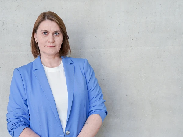 Eine Person mit kurzen braunen Haaren trägt einen blauen Blazer und schaut direkt in die Kamera, vor einer hellgrauen Wand.
