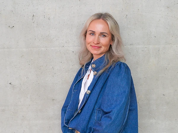 Eine Frau mit blonden Haaren trägt eine blaue Denim-Jacke und steht vor einer neutralen, grauen Wand. Sie lächelt freundlich in die Kamera.