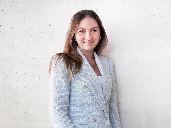 Eine lächelnde Frau mit langen, braunen Haaren trägt einen hellen, zweireihigen Blazer und steht neben einer hellen, strukturierten Wand.