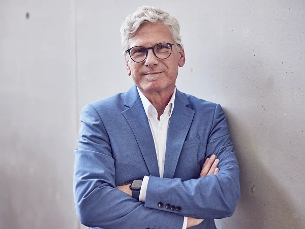 Ein älterer Mann mit grauen, kurzen Haaren und Brille trägt einen blauen Anzug und steht mit verschränkten Armen an einer Betonwand. Er lächelt freundlich in die Kamera.