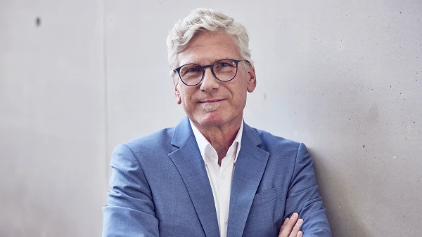 Ein älterer Mann mit grauen, kurzen Haaren und Brille trägt einen blauen Anzug und steht mit verschränkten Armen an einer Betonwand. Er lächelt freundlich in die Kamera.