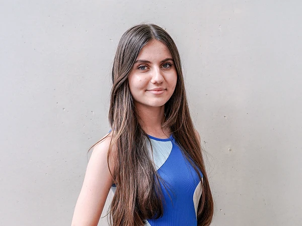 Eine junge Frau mit langen, glatten Haaren steht vor einer grauen Wand und trägt ein ärmelloses, hellblau und dunkelblau gestreiftes Oberteil. Sie lächelt leicht und hat einen entschlossenen Blick.