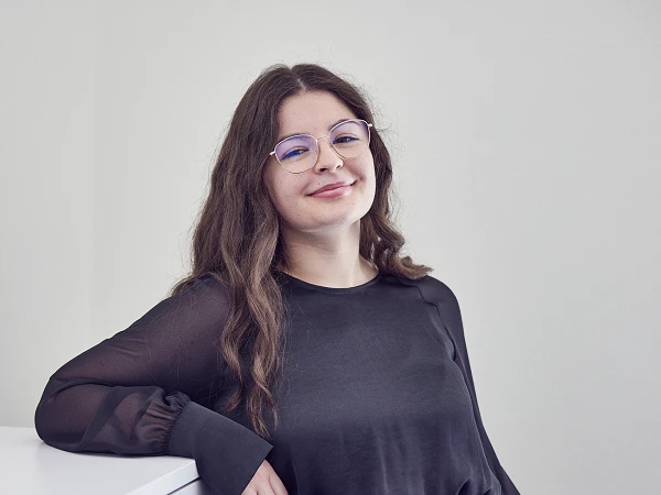 Eine lächelnde Frau mit langen, lockigen Haaren steht an einer weißen Möbelkante und trägt ein schwarzes Oberteil sowie graue Hosen. Sie sieht freundlich in die Kamera und hat eine Brille auf.