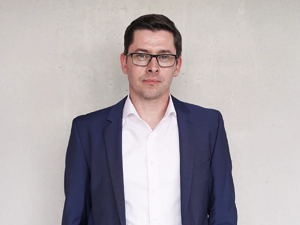 Ein Mann mit Brille trägt einen dunkelblauen Anzug und steht vor einer hellgrauen Wand.