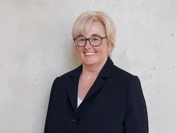 Eine Frau mit blonden, kürzeren Haaren und Brille trägt einen dunkelblauen Blazer und lächelt vor einer neutralen, hellen Wand.