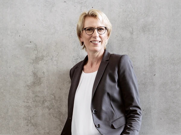 Eine lächelnde Frau mit kurzen blonden Haaren und Brille trägt einen grauen Blazer über einem weißen Oberteil und steht vor einer strukturierten grauen Wand.