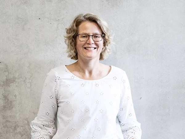 Eine lächelnde Frau mit Locken steht mit den Händen in den Taschen an einer grauen Wand. Sie trägt ein weißes, luftiges Oberteil und hat eine entspannte Ausstrahlung.