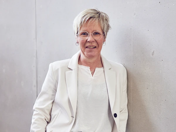 Eine lächelnde Frau mit kurzen, blonden Haaren und Brille steht an einer grauen Wand, gekleidet in einem weißen Oberteil und einem beigen Blazer.