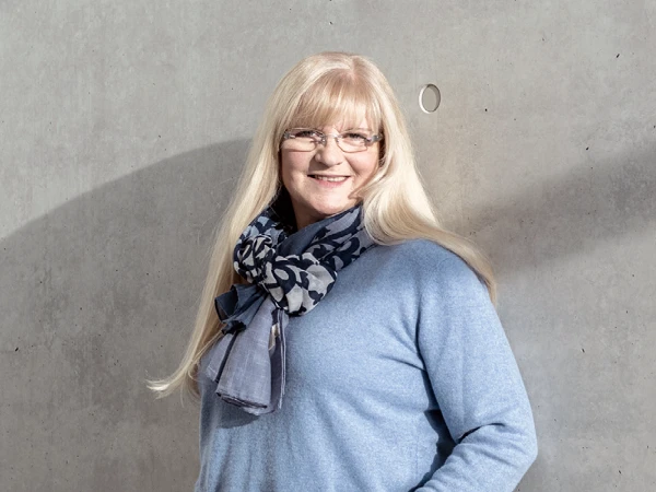 Eine lächelnde Frau mit langen, blonden Haaren und Brille trägt einen blauen Pullover und einen gemusterten Schal. Sie steht an einer grauen Betonwand und hat die Hände in den Taschen.