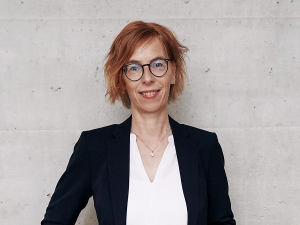 Eine lächelnde Frau mit blonden, lockigen Haaren und Brille steht mit verschränkten Armen vor einer grauen Wand. Sie trägt einen schwarzen Blazer über einer weißen Bluse und wirkt selbstbewusst.