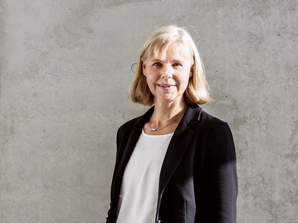 Eine Frau mit blonden, schulterlangen Haaren steht lächelnd vor einer grauen, strukturierten Wand. Sie trägt eine schwarze Jacke und ein weißes Oberteil.