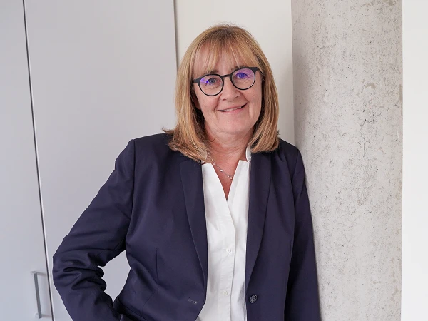 Eine Frau mit blonden Haaren und einer Brille lächelt freundlich, während sie in einem modernen Büro an einer Säule lehnt. Sie trägt einen dunklen Anzug und eine weiße Bluse.