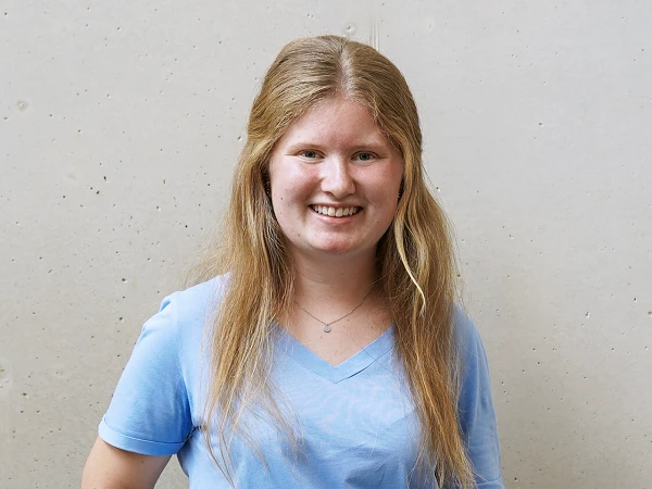 Eine lächelnde junge Frau mit langen, blonden Haaren trägt ein hellblaues T-Shirt und steht vor einer neutralen, grauen Wand.