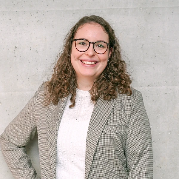 Eine lächelnde Frau mit lockigem Haar steht vor einer grauen Betonwand und trägt ein kariertes Blazer-Jackett über einem weißen Oberteil.